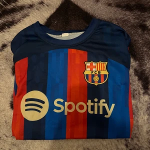 FC Barcelona Lewandowski matchtröja - Snygg FC Barcelona matchtröja med Lewandowski och nummer 9 på ryggen. Tröjan har klassiska blå och röda ränder, klubbmärke på bröstet, La Liga-märke på ärmen och Spotify som sponsor. Tillverkad i lätt och ventilerande material, perfekt för fotboll eller att bära till vardags.
