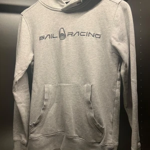 Grå hoodie från Sail Racing - Snygg ljusgrå hoodie från Sail Racing med broderad logga på bröstet. Klassisk känguruficka framtill och huva med dragsko. Perfekt för chill dagar eller när du vill ha en clean och sportig look. Jätte bra skick! Och väldigt snygg hoodie! Nypris är 1500 tror jag! 