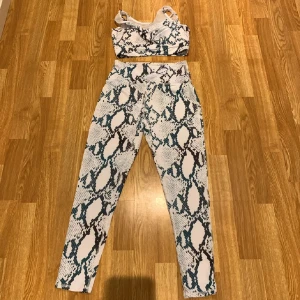 Vit och svart sportig linnetopp med ormmönster - Säljer en snygg sportig linnetopp och tights med ormmönster i vitt, svart och mörkgrönt. Toppen har breda axelband och är tillverkad i stretchigt syntetmaterial som passar perfekt till träning. Mönstret ger en cool och trendig vibe. Tryck ej köp nu