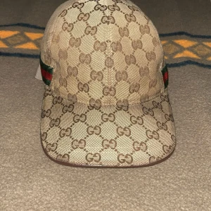 Gucci keps - Snygg Gucci keps i beige och brunt med klassiskt GG-monogram över hela kepsen. Har grönt och rött band på sidan och justerbar rem i brunt läder bak. Tillverkad i canvas och fodrad med mjukt tyg på insidan. Perfekt för dig som vill sticka ut med en lyxig accessoar.