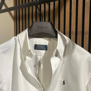 Vit skjorta från Polo Ralph Lauren - Klassisk vit skjorta från Polo Ralph Lauren med button down-krage och broderad marinblå logga på bröstet. Skjortan har lång ärm, knappar framtill och är tillverkad i mjuk bomull. Perfekt för en clean och stilren look. Säljes för att den är för stor på mig.