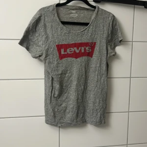 Grå Levi's t-shirt med röd logga - Snygg grå t-shirt från Levi's med klassisk röd logga framtill. T-shirten har rund halsringning och korta ärmar. Perfekt för en avslappnad och cool stil. Tillverkad i mjuk bomull för skön känsla hela dagen.