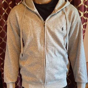 Grå hoodie från Polo Ralph Lauren - Snygg grå hoodie från Polo Ralph Lauren med dragkedja, huva med snörning och klassisk broderad logga på bröstet. Tillverkad i mjuk bomullsmix, perfekt för chill dagar. Fickor framtill och ribbade muddar för extra komfort.Helt ny säljer den så den är i fel storlek!
