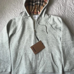 Grå hoodie från Burberry med dragkedja - Snygg ljusgrå hoodie från Burberry med klassiskt rutigt foder i luvan och broderad logga på bröstet.