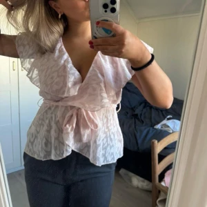 Rosa omlottblus med volang - Supersöt ljusrosa omlottblus med volangdetaljer och knyt i midjan. Blusen är transparent och har leopardmönster. Har använt den endast en gång, i gott skick.💓