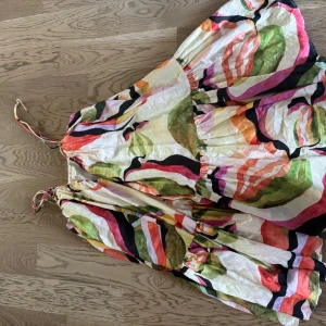 Färgglad volangklänning från H&M - Superfin långklänning från H&M med färgstarkt abstrakt mönster i rosa, orange, grön, svart och vit. Klänningen har tunna axelband, volanger och luftig passform – perfekt för varma dagar och sommarvibbar.