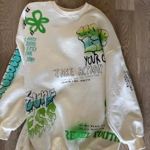 Vit sweatshirt med graffiti från Bershka - Cool vit sweatshirt från Bershka med graffitiinspirerade tryck i grönt, svart och blått. Oversized passform, ribbade muddar och crewneck. Trycken har streetstyle-vibe med texter och sprayade detaljer. Perfekt för dig som gillar unik och edgy stil.