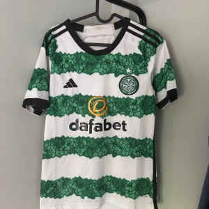 Celtic FC randig fotbollströja Adidas - Säljer en Celtic FC fotbollströja från Adidas med gröna och vita ränder i ett unikt mönster. Tröjan har svart krage och detaljer, klubbmärke på bröstet och sponsortryck framtill. Perfekt för dig som vill sticka ut på planen eller läktaren.