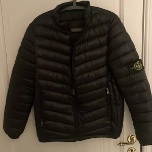 Svart dunjacka från Stone Island - Säljer en svart dunjacka från Stone Island med klassisk patch på ärmen. Jackan har quiltad design, dragkedja framtill och två fickor med dragkedja. Perfekt för kalla dagar och riktigt snygg streetstil.