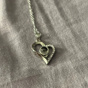 Halsband Med Hjärthänge  - Snyggt silverfärgat halsband med ett hjärthänge dekorerat med små glittrande stenar. I mitten finns en rund sten som projicerar 'Jag älskar dig' på flera språk när man kollar noga på den 🌹
