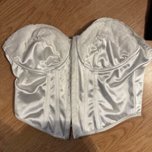 Vit satin korsett med spetsdetaljer - Supersnygg vit korsett i glansig satin med spets på kuporna och hakknäppning framtill. Axelbandslös och croppad modell.