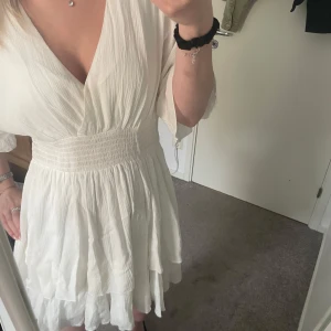 Vit klänning med volang - Supersöt vit kortklänning med omlott och v-ringning. Klänningen har bred smock i midjan, volangig kjol och luftiga korta ärmar. Perfekt för sommarens alla tillfällen och ger en fräsch och luftig känsla.