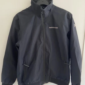 Vindjacka Peak Performance - Snygg marinblå vindjacka från Peak Performance med hög krage och dragkedja framtill. Jackan har två fickor med dragkedja och diskret logga på bröstet och ärmen. Perfekt för blåsiga dagar och enkel att matcha med allt. Jackan har inga defekter och är inprincip som ny! Passar perfekt till sena sommarkvällar eller tidig höst! 