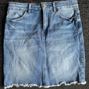 Blå jeanskjol med fransig kant - Snygg blå jeanskjol i klassisk modell med 4 fickor och fransig, rå kant nertill. Kjolen har bälteshällor och dragkedja med knapp framtill.