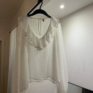 Vit blus med volang från NLY Trend - Supersöt vit blus från NLY Trend med diskret prickigt mönster och volangdetalj längs v-ringningen. Blusen har långa, luftiga ärmar och knäppning i nacken. Perfekt för dig som gillar romantisk och trendig stil.