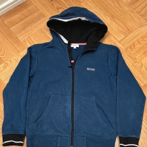 Blå zip-hoodie från BOSS med huva - Mörkblå hoodie från BOSS med dragkedja, huva och vita detaljer på muddar. Jackan har fickor framtill och broderad logga på bröstet. Tillverkad i bomull och polyester för en skön och avslappnad känsla.