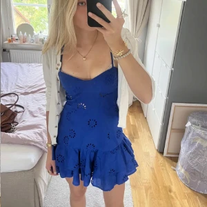 Blå kort klänning med volang - Supersöt blå kort klänning med smala axelband, volangkant och broderade hålmönster. Klänningen har en figurnära passform upptill och en utställd kjol med dubbla volanger. Perfekt för sommarens alla tillfällen!