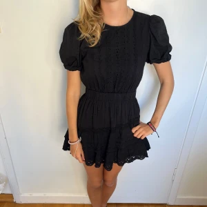 Svart kort klänning från Zara  - Supersöt svart kort klänning från Zara med puffärmar, broderade detaljer och volangkant nertill. Klänningen har markerad midja och rund halsringning, perfekt för dig som gillar en feminin och trendig stil.