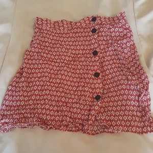 Rödvit mönstrad minikjol med knappar - Superfin röd kortkjol med vitt mönster och svarta knappar framtill. Kjolen är i 100% viskos och har en lätt A-linjeformad passning. Perfekt för varma dagar när du vill ha något luftigt och snyggt.