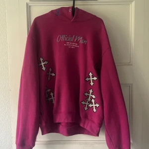 Cerise hoodie från Official Man - Säljer en cerise hoodie från Official Man med broderade korsdetaljer i vitt och svart på framsidan. Tröjan har tryckt text på bröstet och broderad text på huvan. Oversized passform och mjukt material, perfekt för dig som gillar streetwear.