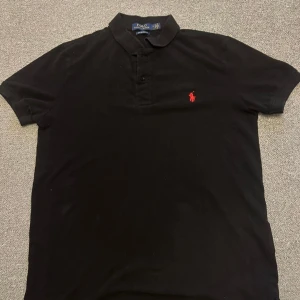 Svart pikétröja Ralph Lauren S - Svart pikétröja från Ralph Lauren med klassisk krage och knappar framtill. Röd broderad logga på bröstet och slim fit-passform. Tillverkad i bomull, perfekt för en clean och stilren look.
