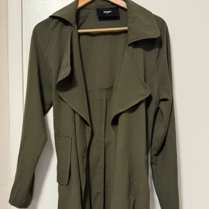 Olivgrön trenchcoat från BikBok XS - Snygg olivgrön trenchcoat från BikBok i storlek XS. Jackan har långa ärmar, bred krage och knytskärp i midjan för en clean och stilren look. Praktiska fickor framtill. Lätt material som passar till många outfits.