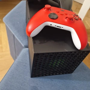 Xbox series x 1 tb - Bilderna visar en Xbox Series X spelkonsol med två handkontroller, en svart och en röd. Detta är ingen bok, utan en spelkonsol för gaming och underhållning.