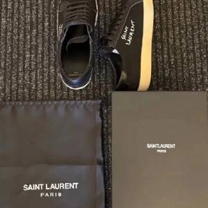 Svarta Saint Laurent sneakers - Snygga svarta sneakers från Saint Laurent med vit logga broderad på sidan. Skorna har svart snörning, ovandel i canvas och detaljer i skinn. Sulan är beige och ger en clean kontrast. Perfekta för dig som gillar stilrena och exklusiva sneakers. Äkta kvitto finns