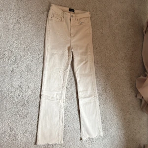 Beige raka jeans med råa kanter från Vero Moda  - Snygga beige jeans från Vero Moda med raka ben och råa, fransiga benslut. Klassisk femficksmodell med knapp och dragkedja framtill. Perfekta för en avslappnad och trendig look.