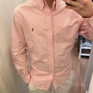 Polo Ralph Lauren (Rosa skjorta) - Säljer en klassisk rosa skjorta från Polo Ralph Lauren med button-down krage och broderad logga på bröstet. Skjortan är oanvänd och redo för en ny ägare! Perfekt för en clean och stilren look. Den är storlek S!