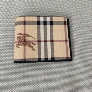 Burberry plånbok - Snygg plånbok från Burberry i klassiskt beige rutigt mönster med brunt ryttaremblem på framsidan. Insidan är svart skinn med flera kortfack och fönster för ID. Perfekt accessoar för dig som gillar stilrena detaljer. Pris 300kr