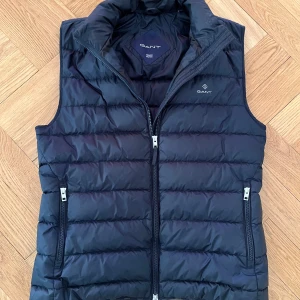 Marinblå dunväst från GANT strl M - Snygg marinblå dunväst från GANT med quiltad design och dragkedja framtill. Västen har två fickor med dragkedja och hög krage. Perfekt att bära över en tröja för extra värme.