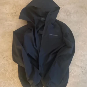 Svart vindjacka från Peak Performance - Säljer en svart vindjacka från Peak Performance med huva och dragkedja. Jackan har en stilren design och är perfekt för blåsiga dagar. Logotypen syns diskret på bröstet. Passar dig som gillar funktionella och snygga ytterplagg.