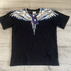 Svart t-shirt med blått och vitt vingmönster från Marcelo Burlon - OBS: storlek S men passar som M Säljer en svart t-shirt från Marcelo Burlon med ett stort, detaljerat vingmönster i vitt och blått över axlar och bröst. T-shirten har rund hals och korta ärmar. Perfekt statement-plagg för dig som gillar unika designer.