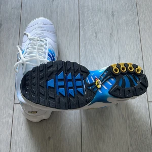 Nike Air Max Plus vita och blå sneakers - Nike Air Max Plus sneakers i vitt och blått med svarta och gula detaljer. Skorna har en vågig design på ovandelen, synliga Air Max-enheter i sulan och snörning framtill. Perfekta för dig som vill sticka ut med en klassisk och sportig look.
