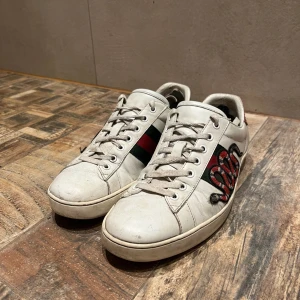 Gucci ace  - Säljer ett par vita sneakers från Gucci med klassiska gröna och röda ränder på sidorna samt en broderad röd och svart orm. Skorna är i okej skick. Defekterna på skorna är att högra skon är lite creasad, sen är det ett litet hål i bak på sulan, och den sista defekten är att skinnet inuti skorna har hälarna har gått upp, men man tänker inte på det och det känns inte heller. Där av det låga priset. Jag är öppen för byten!