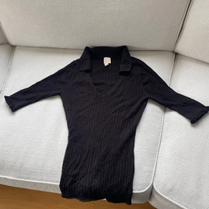 Svart ribbad topp - Säljer en svart ribbad topp med v-ringning och trekvartsärm. Tröjan har en klassisk krage och är figurnära i modellen. Perfekt att styla till både jeans och kjol.