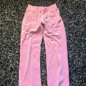 Rosa velourbyxor från Juicy Couture - Säljer ett par klassiska rosa velourbyxor från Juicy Couture med broderad JC-logga på fickan framtill och fickor med knappar bak. Byxorna har normal passform och dragsko i midjan. Perfekta för en avslappnad och trendig look.