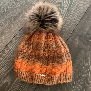 Orange och brun stickad mössa med pälsboll - Snygg stickad mössa i orange och bruna nyanser med stor, fluffig pälsboll på toppen. Mössan har ett diskret metallmärke framtill och ett trendigt kabelstickat mönster. Perfekt för att hålla värmen och ge en färgklick till din outfit.