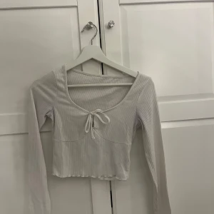 Vit ribbad croppad topp med knyt - Säljer en vit ribbad croppad topp med lång ärm och söt knytning framtill. Toppen har en  rundad ringning och är perfekt för en trendig vardagslook.