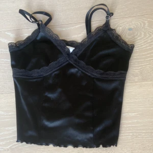 Svart sammetslinne med spets från H&M Divided - Säljer ett svart sammetslinne från H&M Divided med tunna axelband och fina spetsdetaljer längs urringningen och upptill. Linnet har en croppad passform och vågig nederkant. Perfekt för att lyfta vilken outfit som helst!