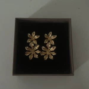 Guldiga blomformade örhängen - Säljer ett par guldiga örhängen i form av blommor. De har en elegant design med två sammankopplade blommor per örhänge och sitter med plupp baktill. Använda en gång.