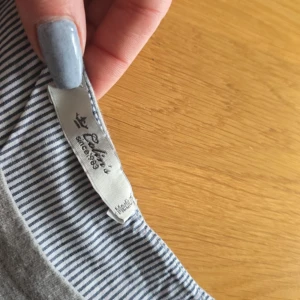 Grå långärmad tröja med ränder och text från Colin's😍 - Säljer en grå långärmad tröja från Colin's med texten 'GORGEOUS' framtill. Nedre delen och insidan av kragen har smala blåvita ränder och knappar baktill. Perfekt för en avslappnad stil.