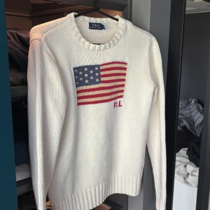 Vit stickad tröja från Polo Ralph Lauren - Vit stickad tröja från Polo Ralph Lauren med amerikansk flagga och RL-broderi på bröstet. Rund halsringning och ribbade muddar vid ärmslut och nederkant. Klassisk och stilren design.