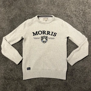 Ljusgrå stickad tröja från Morris - Säljer en ljusgrå stickad tröja från Morris med broderad logotyp och emblem på bröstet. Tröjan har rund halsringning med diskret randig detalj och ribbade muddar vid ärmslut och nederkant. Modellen är 169cm och väger 59✅