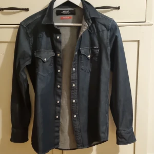 Mörkblå jeansskjorta från Replay - Säljer en snygg mörkblå jeansskjorta från Replay Hyperflex med tryckknappar och två bröstfickor. Skjortan har klassisk krage och lång ärm, perfekt för en cool och avslappnad stil. Skriv vid minsta fundering!