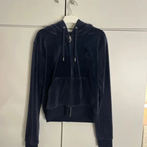 Mörkblå velour hoodie från Juicy Couture - Säljer en klassisk mörkblå hoodie från Juicy Couture i mjuk velour. Tröjan har dragkedja med J-detalj, huva och stora fickor framtill. Perfekt för en avslappnad och trendig look.