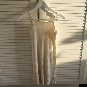 Ljus ribbad kortklänning med smala axelband - Säljer en ljus ribbad kortklänning med smala axelband och rak halsringning. Klänningen har en enkel och stilren design, perfekt för varma dagar. Materialet är stretchigt och följsamt.