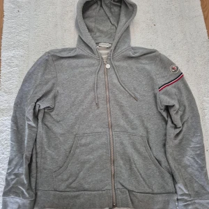 Moncler Maglia Cardigan Zip Hoodie L - Säljer en fet hoodie med dragkedja och huva som är i väldigt bra skick!. Tröjan har fickor framtill och en broderad Moncler-logga på ärmen tillsammans med en randig detalj i rött, vitt och blått. Passa på att fynda denna hoodie och stick ut bland mängden! Liiten i storleken så den passar en M.