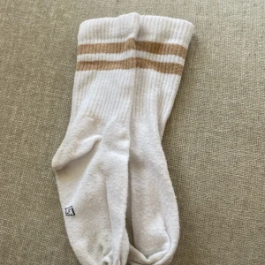 Vita sportiga strumpor med bruna ränder storlek barn - Säljer ett par vita ribbade strumpor med två bruna ränder upptill. Strumporna är höga och har en sportig retrostil, perfekta till sneakers eller träning. Mjukt material och bekväm passform. Barn 